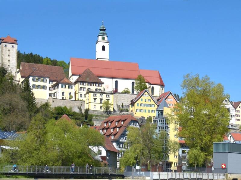 Eine malerische Hügellandschaft mit bunten Fachwerkhäusern, über der eine Kirche mit rotem Dach thront, unter einem blauen Himmel mit einigen Wolken.
