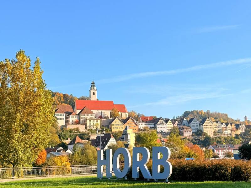 Eine hügelige Stadtlandschaft mit traditionellen Häusern, einem Kirchturm und dem Schriftzug "HORB" im Vordergrund.