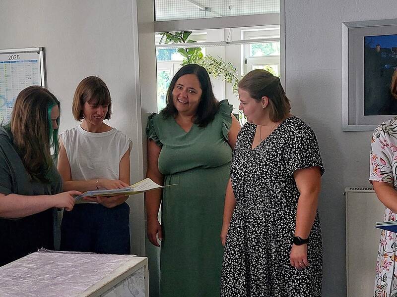 Sieben Frauen in einem Büro stehen zusammen, zwei halten Ordner, ein kleiner Hund sitzt auf dem Boden in der Mitte.