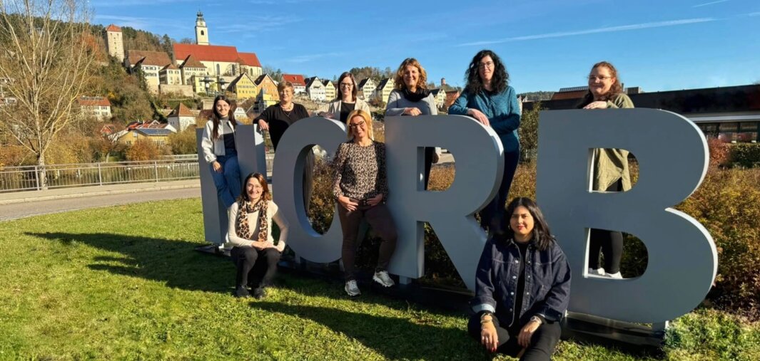 Gruppe von Menschen sitzt und steht um große Buchstaben H, O, R und B auf einer Wiese mit Dorf und Kirche im Hintergrund.