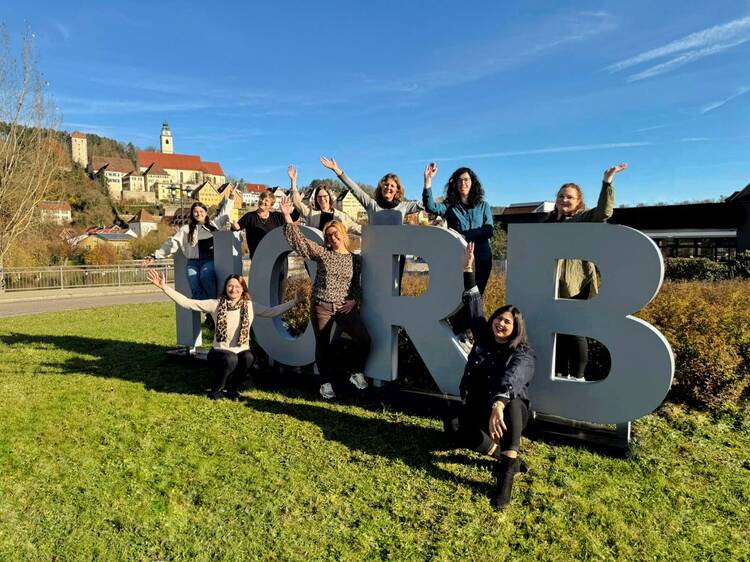 Gruppe von Menschen posiert fröhlich um große graue Buchstaben "H O R B" auf einer Wiese, im Hintergrund Häuser und blauer Himmel.