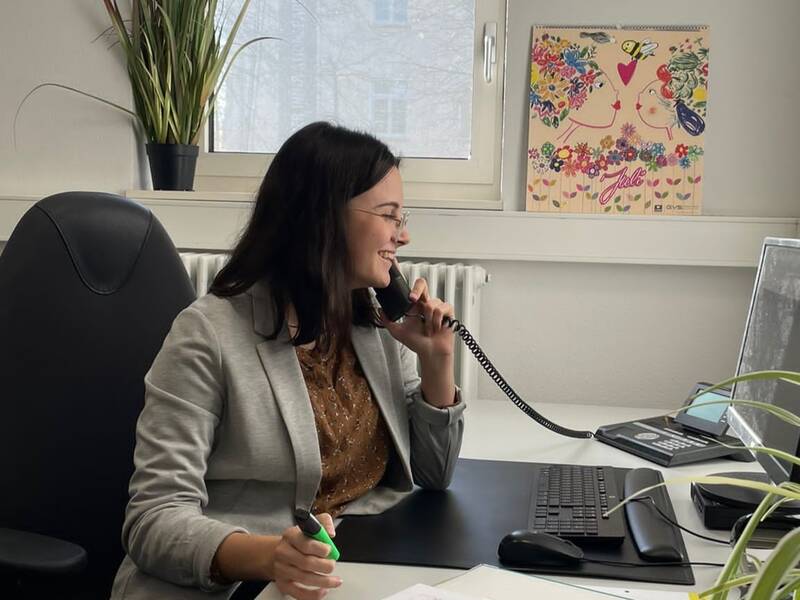 Eine Frau sitzt in einem Büro am Schreibtisch, telefoniert lächelnd und hält einen Stift in der Hand, während ein Computerbildschirm und ein Ordner vor ihr liegen.