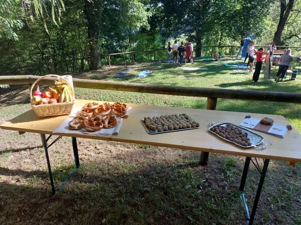 Tisch mit Korb voller Obst und Tellern mit Brezeln und Pralinen im Freien, im Hintergrund Menschen auf einer Wiese unter Bäumen.