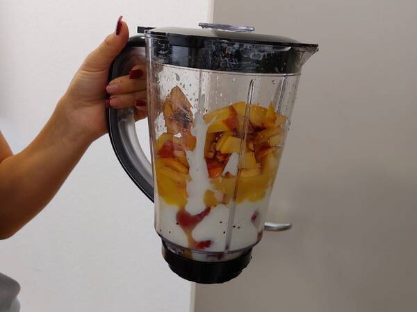 Hand hält Mixbehälter mit Deckel, gefüllt mit Joghurt und Obststücken, vor weißer Wand.