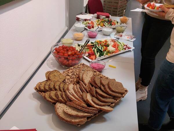 Tisch mit mehreren Brotsorten, Schalen mit Kirschtomaten, Gemüsesticks und verschiedenen Dips, im Hintergrund Personen beim Buffet.