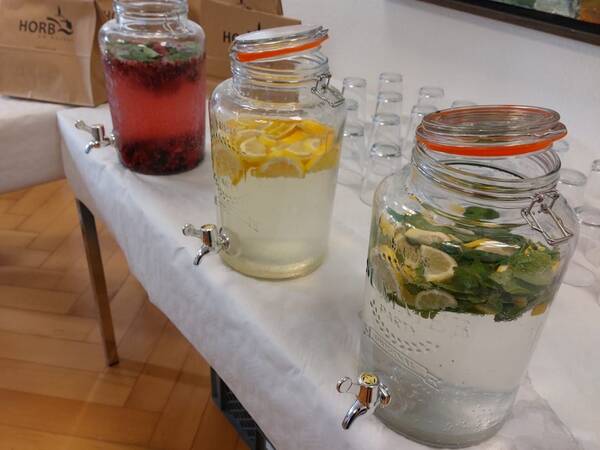 Drei große Glasbehälter mit Zapfhähnen, gefüllt mit Wasser und verschiedenen Früchten und Kräutern, auf einem Tisch mit Papiertüten und Gläsern im Hintergrund.
