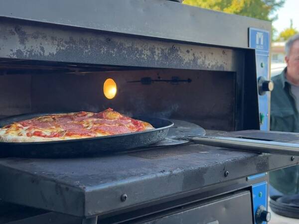 Pizza in schwarzer Backform wird aus einem mobilen Pizzaofen mit Metallgriff geschoben, im Hintergrund unscharfe Bäume und Person.