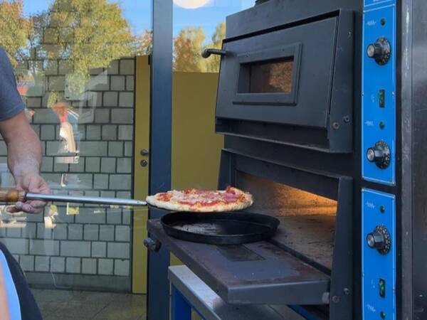 Pizza wird mit einem Pizzaschieber aus einem schwarzen Pizzaofen mit blauer Front geholt.