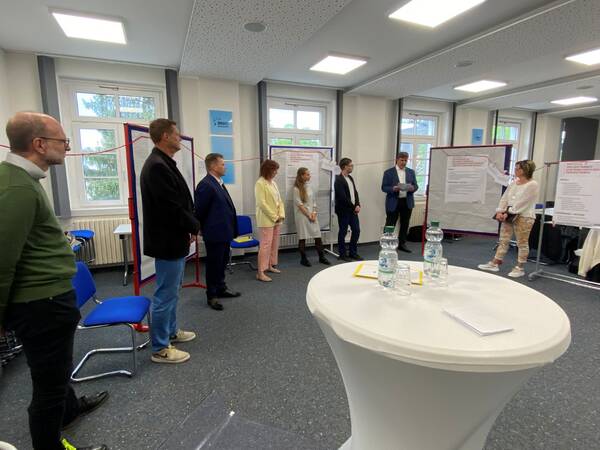 Mehrere Personen stehen in einem Halbkreis in einem hellen Raum mit großen Fenstern, einige blicken auf eine Person, die vor Flipcharts spricht.