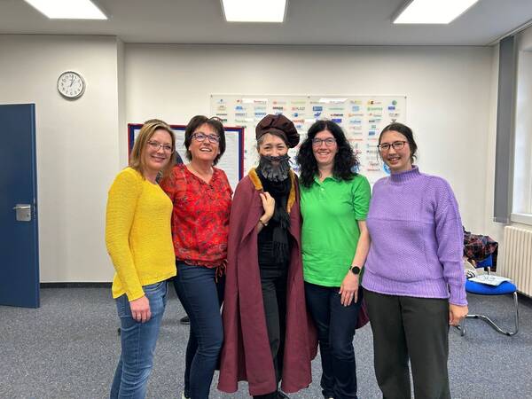 Fünf Frauen stehen nebeneinander in einem Raum mit Uhr und Poster an der Wand. Sie tragen bunte Pullover und eine trägt einen langen Mantel und eine Mütze.
