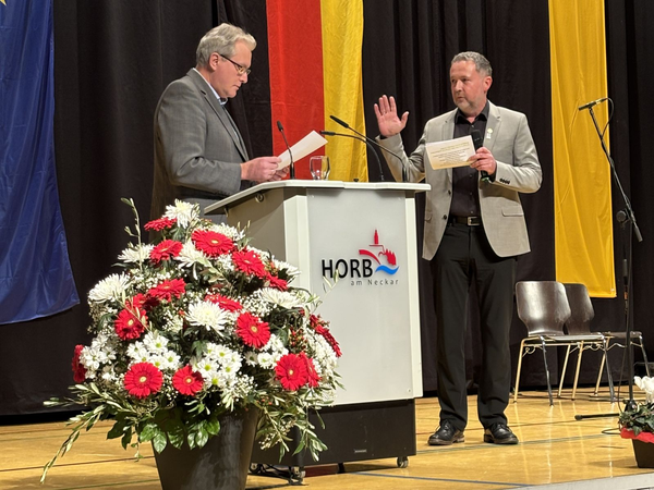 Zwei Männer stehen auf einer Bühne vor einem Rednerpult mit dem Schriftzug 'HORB am Neckar', einer hebt die rechte Hand, der andere hält ein Blatt Papier. Im Vordergrund ein großer Blumenstrauß mit roten und weißen Blumen. Im Hintergrund hängen drei Fahnen in Blau, Rot-Gelb und Gelb.