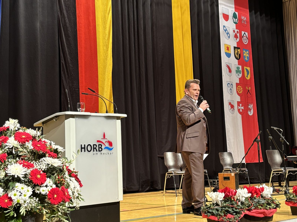 Mann in braunem Anzug spricht mit Mikrofon auf Bühne, links Rednerpult mit Schriftzug 'HORB am Neckar', im Hintergrund bunte Fahnen und Stuhlreihe, vorne Blumenarrangements.