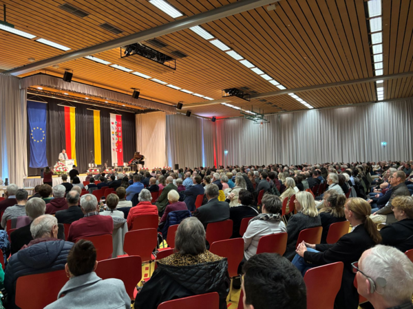 Großer Saal mit Publikum auf roten Stühlen, Bühne mit Rednerpult, Klavier und EU-, Deutschland- sowie weiteren Flaggen.