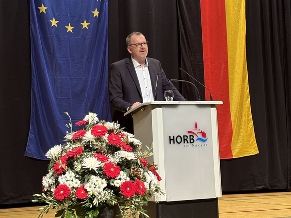 Mann steht hinter einem Rednerpult mit der Aufschrift 'HORB am Neckar', davor ein großer Blumenstrauß mit roten und weißen Blumen, im Hintergrund EU- und Deutschlandflagge.