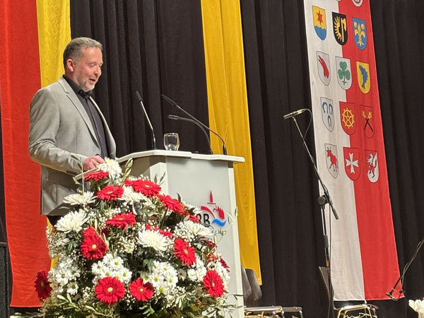 Mann in grauem Sakko steht am Rednerpult mit Mikrofonen, davor ein großer Blumenstrauß mit roten und weißen Blumen, im Hintergrund bunte Fahnen mit Wappen.