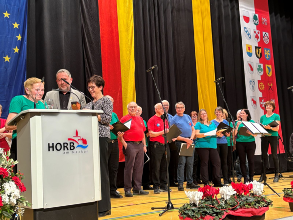 Mehrere Personen stehen auf einer Bühne vor einem Rednerpult mit der Aufschrift 'HORB am Neckar'. Im Hintergrund hängen große Fahnen in den Farben Blau, Rot, Gelb und Schwarz sowie eine Tafel mit Wappen.