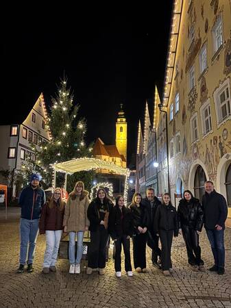 Gruppe von Menschen steht auf gepflastertem Platz vor weihnachtlich geschmücktem Baum und beleuchtetem Pavillon in einer Altstadt bei Nacht