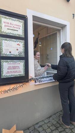 Junge Frau in schwarzer Jacke reicht an einem Fenster einem Menschen im Innenraum einen Becher, neben dem Fenster hängen zwei Schilder mit Texten zu einer Spendenaktion.