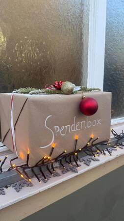 Mit braunem Papier umwickelte Spendenbox mit Tannenzweig, goldener Kugel, roter Christbaumkugel und Lichterkette auf Fensterbank