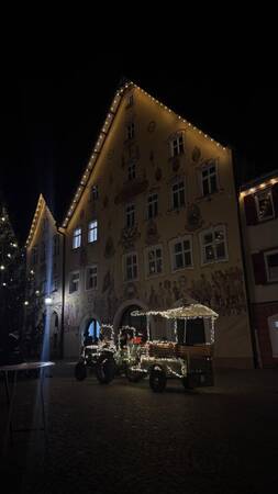 Beleuchtetes historisches Gebäude mit bemalter Fassade und festlich geschmücktem Traktor mit Anhänger davor in der Nacht