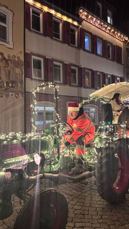 Person in roter Arbeitskleidung und Weihnachtsmütze sitzt auf mit Lichterketten geschmücktem Traktor vor einem beleuchteten Gebäude