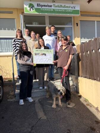 Gruppe von Menschen steht vor dem Eingang eines Gebäudes mit Schild 'Allgemeiner Tierhilfsdienst e.V. Tierheim "Renate Lang"', eine Person hält einen großen Spendenscheck, eine weitere Person hält eine Hundeleine mit einem Hund.