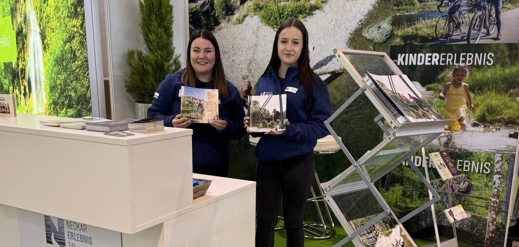 Zwei Personen stehen an einem Messestand mit Broschüren. Im Hintergrund sind grüne Bäume und ein Weg zu sehen. Auf dem Tisch steht ein Logo mit 'Neckar Erlebnis Tal'.