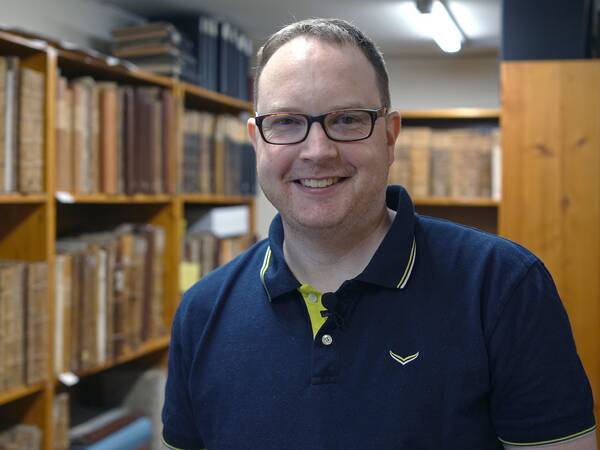 Mann mit Polohemd steht in einer Bibliothek mit Regalen voller alter Bücher. Die Regale sind aus Holz und die Bücher haben gebundene Einbände.