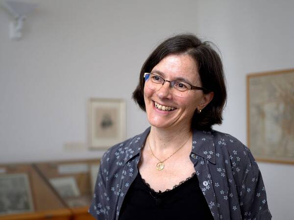 Frau mit schulterlangen, dunklen Haaren, trägt eine graue Bluse mit weißen Mustern und eine goldene Halskette. Im Hintergrund sind Bilderrahmen und eine Vitrine sichtbar.