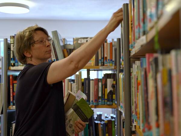 Eine Person mit kurzen blonden Haaren greift nach einem Buch in einem Bücherregal. Sie hält mehrere Bücher in der anderen Hand. Im Hintergrund sind zahlreiche Bücher sichtbar.