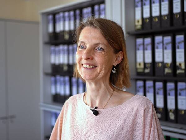 Frau mit kurzen braunen Haaren, trägt rosa Bluse und schwarzen Ansteckmikrofon, steht vor Regal mit Ordnern.