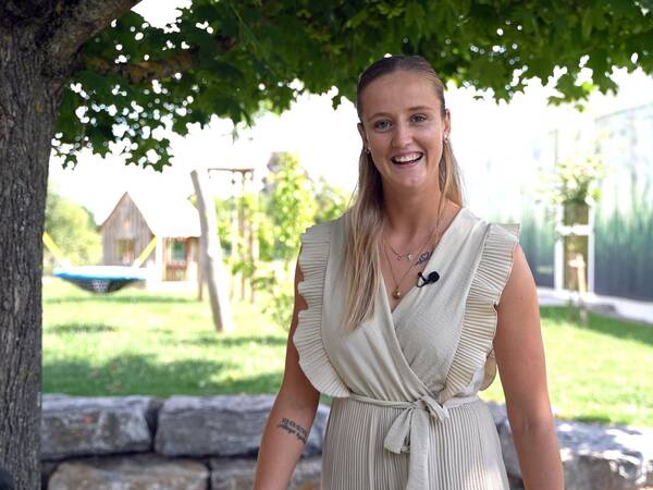 Frau mit langem, glattem Haar trägt ärmelloses, helles Kleid mit Rüschen und mehreren Ketten, steht unter einem Baum im Garten.