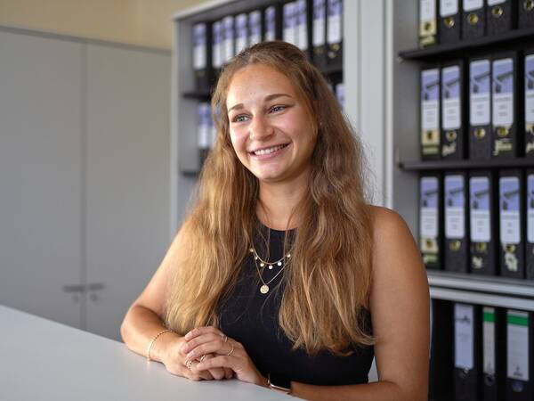 Frau mit langen, braunen Haaren, trägt ein schwarzes Top. Sie steht an einem Tisch vor einem Regal mit Aktenordnern.