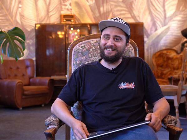 Mann mit schwarzem T-Shirt und Baseballcap sitzt auf einem Sessel in einem Raum mit Pflanzenmotiven. Im Hintergrund sind eine Couch und ein Holzschrank sichtbar.