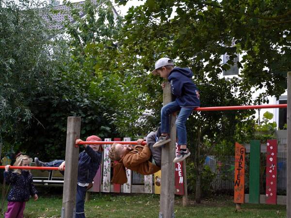 Zwei Kinder spielen an einem Spielgerät im Kindergarten. Ein Kind klettert auf eine Stange, während ein anderes daran hängt. Im Hintergrund sind bemalte Holzwände und Bäume sichtbar.