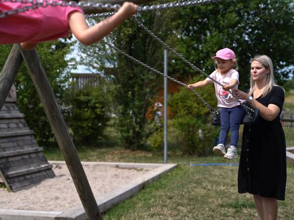 Eine Frau in schwarzem Kleid schiebt ein Kind auf einer Schaukel. Im Hintergrund ist ein Spielplatz mit einem Holzspielhaus und Bäumen zu sehen.