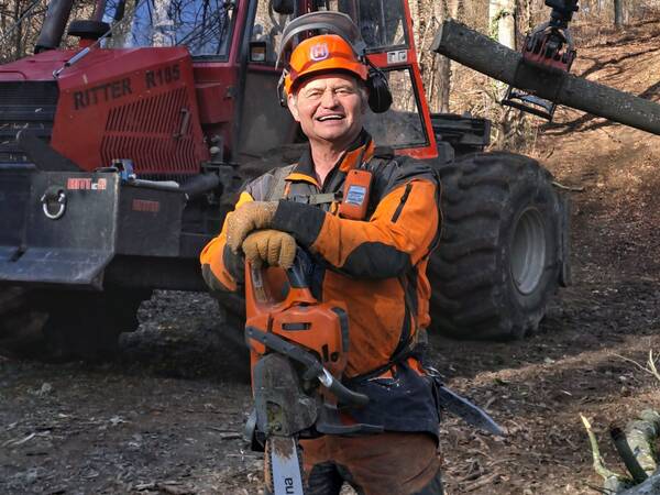 Mann in orange-schwarzer Arbeitskleidung mit einer Motorsäge in der Hand. Im Hintergrund ein roter Holzerntefahrzeug und ein gefällter Baumstamm.