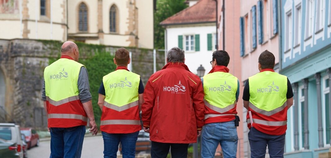 Fünf Personen stehen mit dem Rücken zur Kamera auf einem gepflasterten Platz in Horb am Neckar, tragen gelbe Warnwesten und rote Jacken mit dem Schriftzug 'HORB'