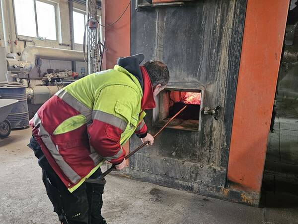 Ein Mann in einer rot-grünen Arbeitsjacke und schwarzer Hose steht vor einem großen Ofen. Er hält einen langen Stab in der Hand und schaut in den Ofen, aus dem rotes Licht strahlt.