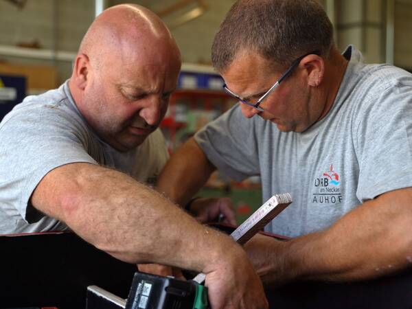Zwei Handwerker in grauen T-Shirts arbeiten zusammen an einem Projekt, halten Werkzeuge und messen Materialien.
