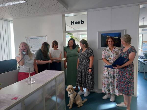 Eine Gruppe von sieben Personen steht in einem Büro. Eine Person am Schreibtisch telefoniert. Ein Hund sitzt auf dem Boden. An der Wand hängt ein Bild und ein Schild mit der Aufschrift 'Horb'.