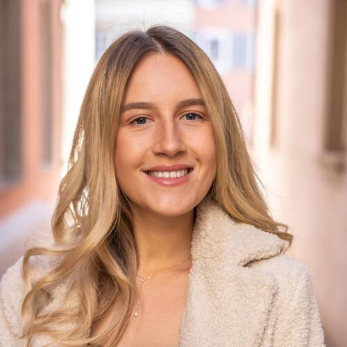 Eine lächelnde Person mit langen blonden Haaren trägt einen hellen Mantel, ein schwarzes Oberteil und Jeans, während sie in einem schmalen, lichtdurchfluteten Durchgang steht.