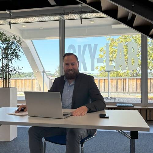 Mann in Businesskleidung sitzt an einem weißen Tisch mit Laptop in modernem Büro mit großen Fenstern und Pflanzen