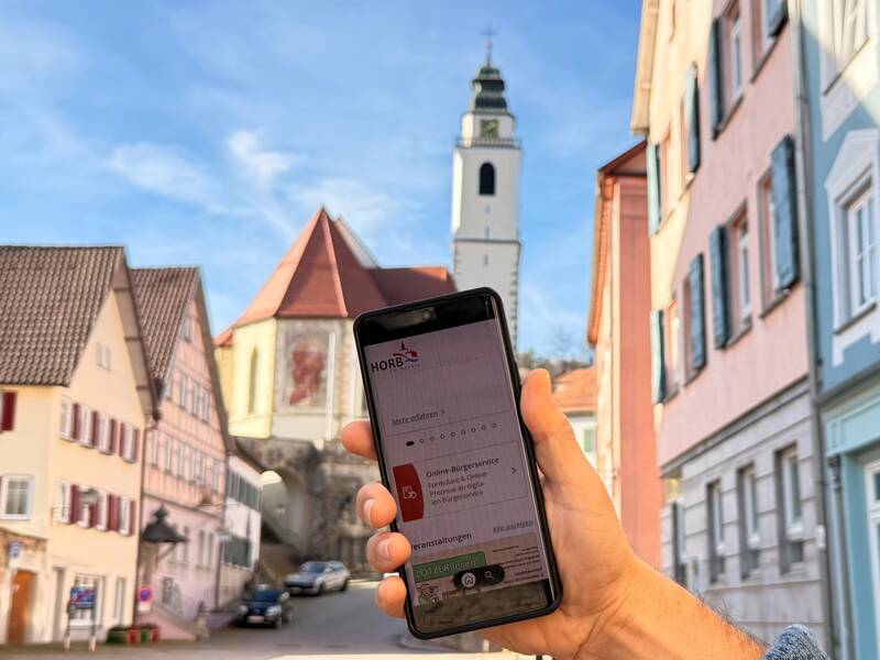 Ein Smartphone wird hochgehalten vor einer malerischen Altstadt-Kulisse mit Fachwerkhäusern und einem Kirchturm im Hintergrund.