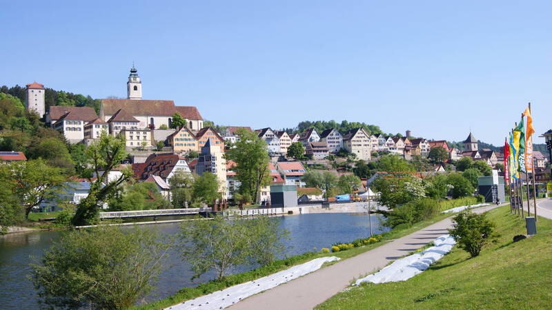 Die Aufnahme zeigt Horb am Neckar mit historischen Gebäuden auf einem Hügel, einem Fluss im Vordergrund und farbenfrohen Flaggen entlang eines Fußwegs.
