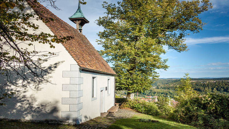 Die Aufnahme zeigt eine kleine weiße Kapelle mit einem roten Ziegeldach und Turm neben einem großen Baum, umgeben von einer idyllischen Landschaft im Hintergrund.