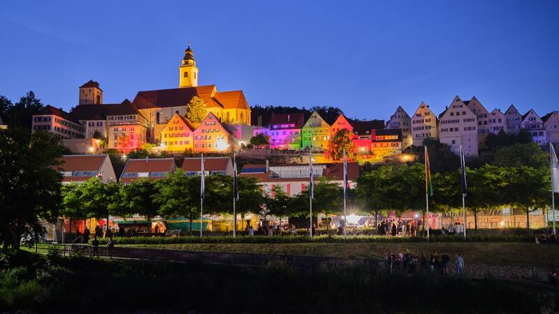 Die historischen Gebäude auf einem Hügel sind nachts in verschiedenen leuchtenden Farben beleuchtet, und im Vordergrund sind Menschen unter Bäumen zu sehen.