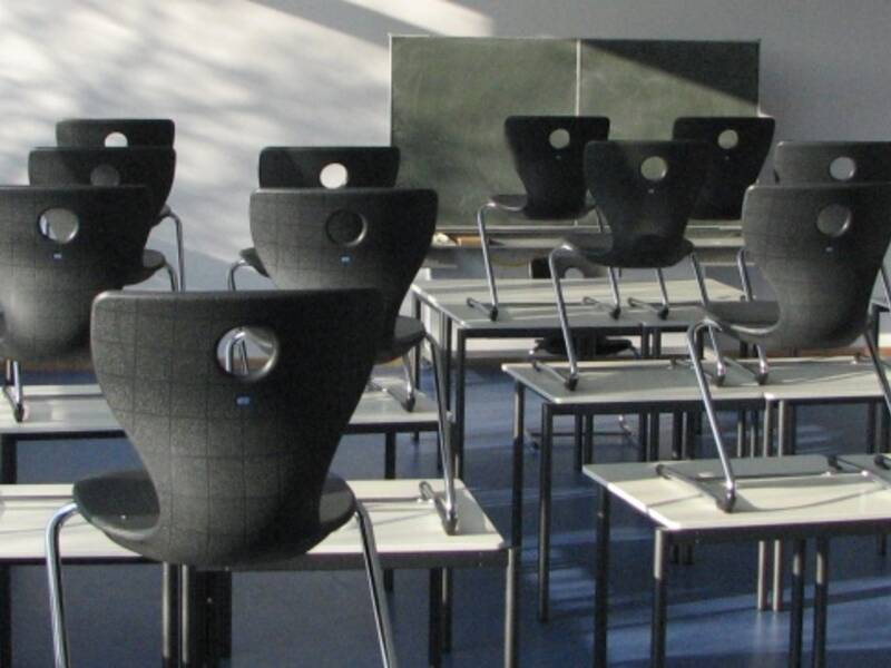 Leerer Klassenraum mit schwarzen Stühlen und Tischen, Sonnenlicht fällt durch große Fenster.