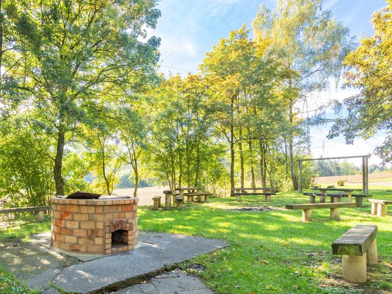 Grüner Park mit mehreren Holzbänken und einem gemauerten Grillplatz unter Bäumen und zwei Schaukeln.