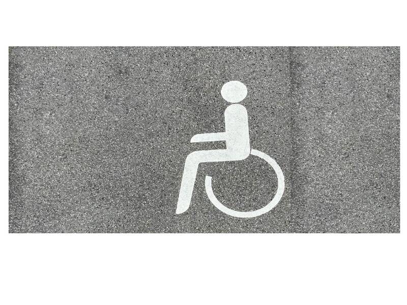 Weißes Rollstuhlsymbol auf grauem Asphalt markiert barrierefreien Bereich.